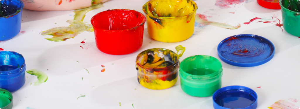 Bunte Dosen mit Fingerfarben auf einem weißen Tisch, umgeben von Farbklecksen.